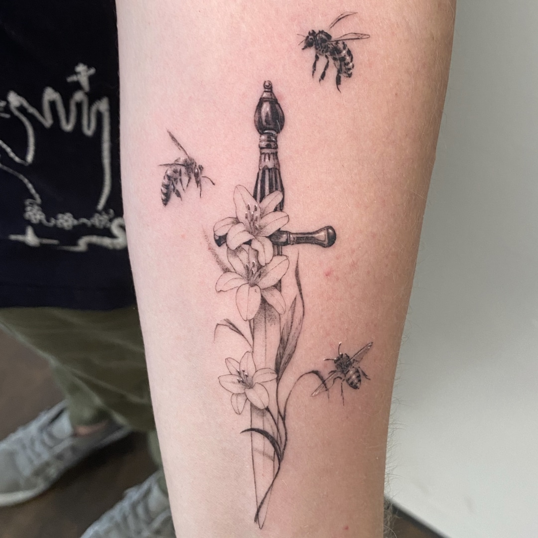 Tattoo of a sword surrounded by flowers and bees.